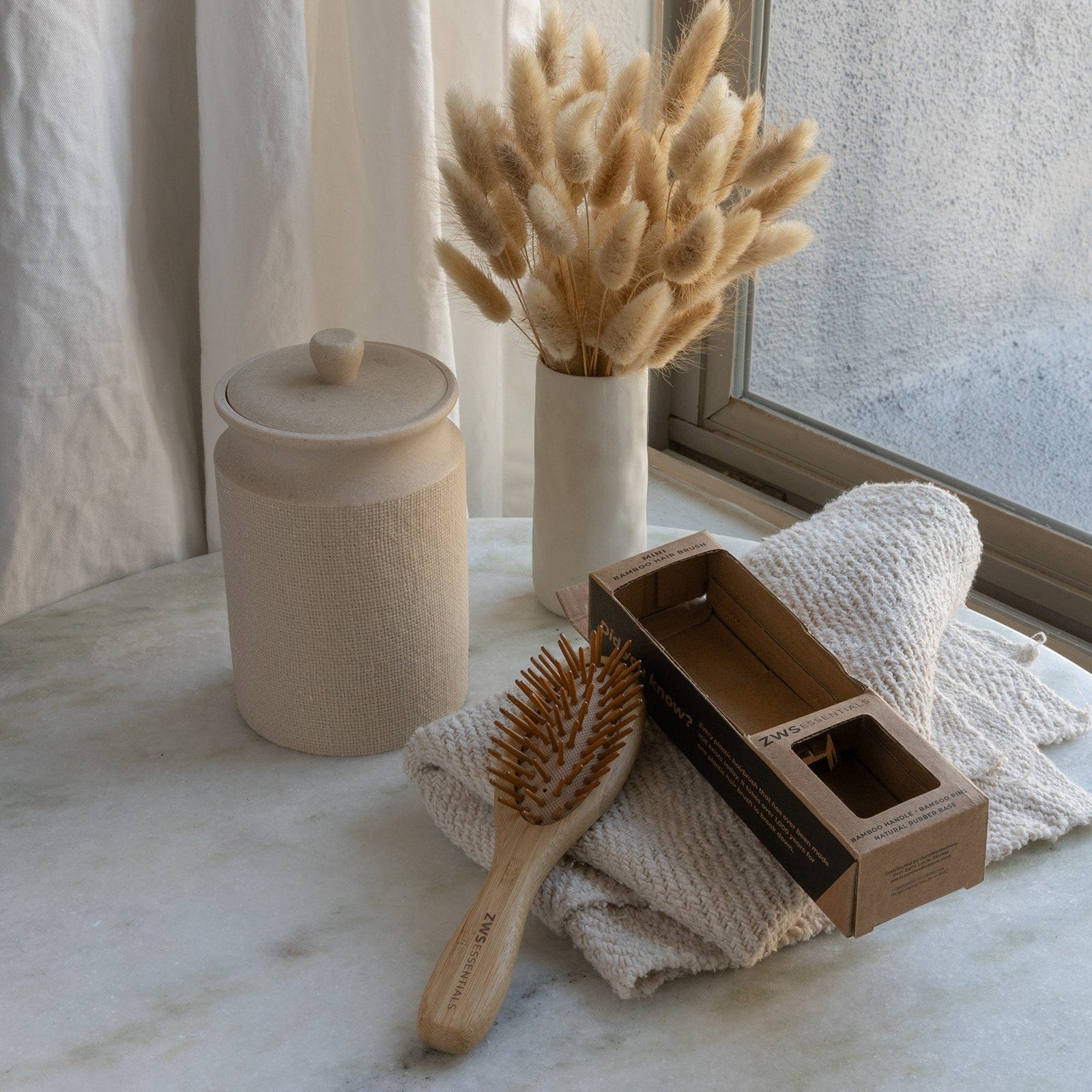 The Mini Bamboo Hair Brush by ZeroWasteStore.com, its box, soft folded towels, a ceramic jar, and a vase with dried beige grasses are neatly arranged on marble near a window with sheer curtains.
