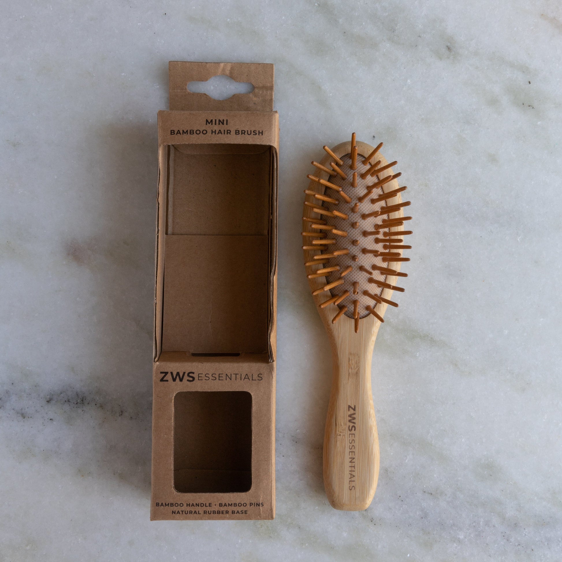 The ZeroWasteStore.com Mini Bamboo Hair Brush—100% bamboo, plastic free, compostable—rests beside its open cardboard box labeled “Mini Bamboo Hair Brush” on a light marble surface.