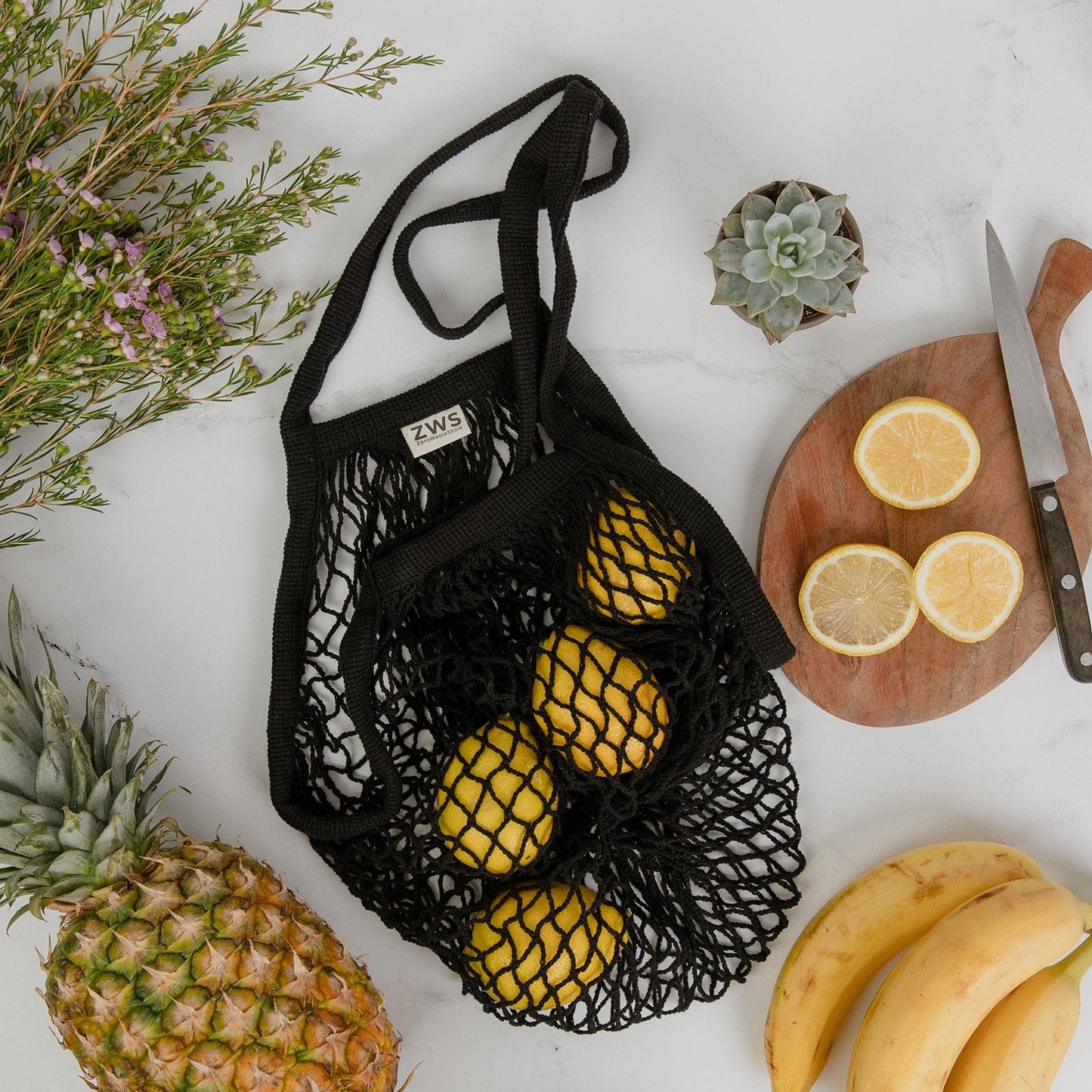 The Organic Cotton String Bag by ZeroWasteStore.com sits on a marble surface with lemons, a pineapple, bananas, a board of sliced lemons, a knife, a succulent plant, and greenery.