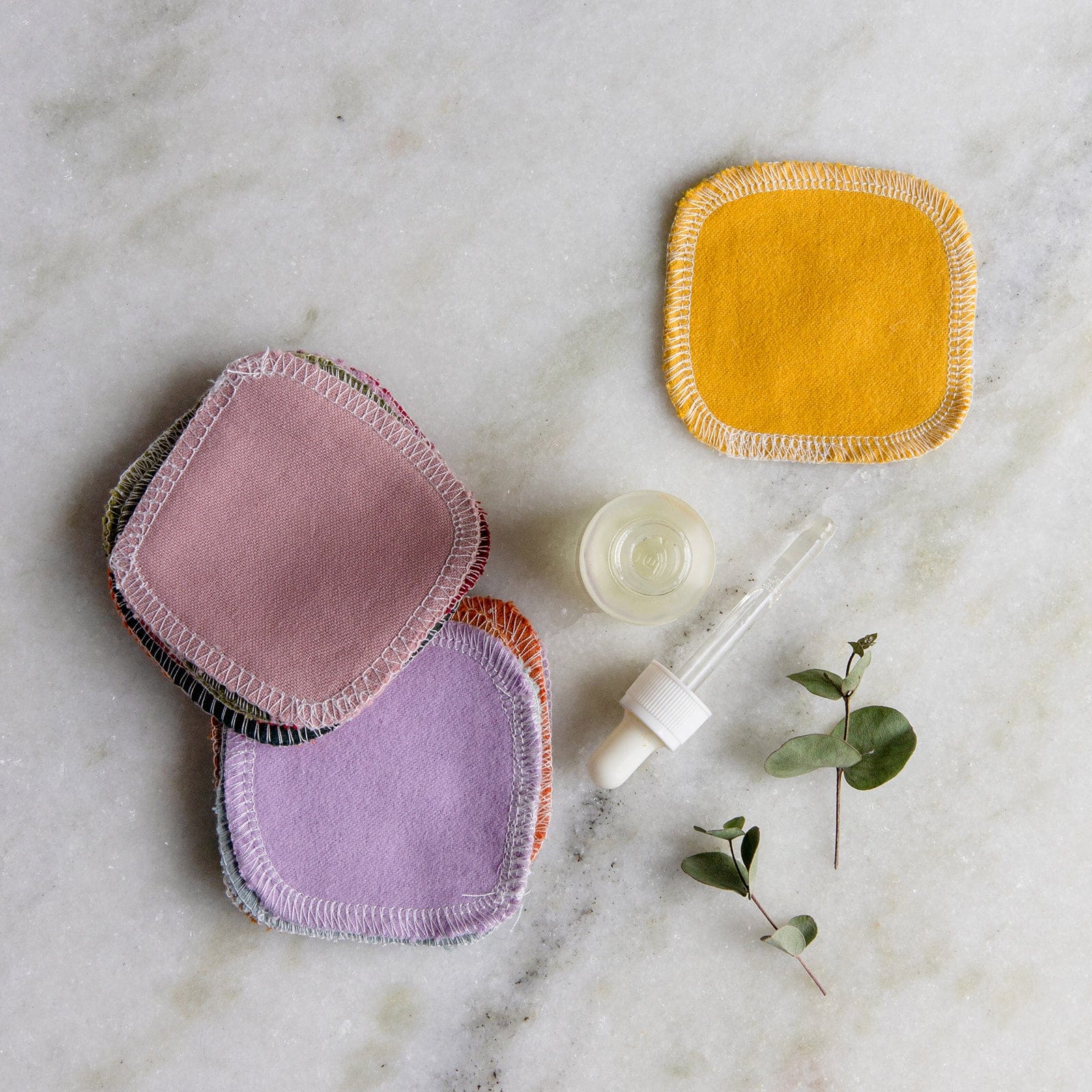Four pastel Reusable Cotton Rounds from ZeroWasteStore.com are stacked on a marble surface beside a yellow organic cotton pad, glass dropper bottle, and sprigs of greenery—ideal for your zero waste skincare routine.