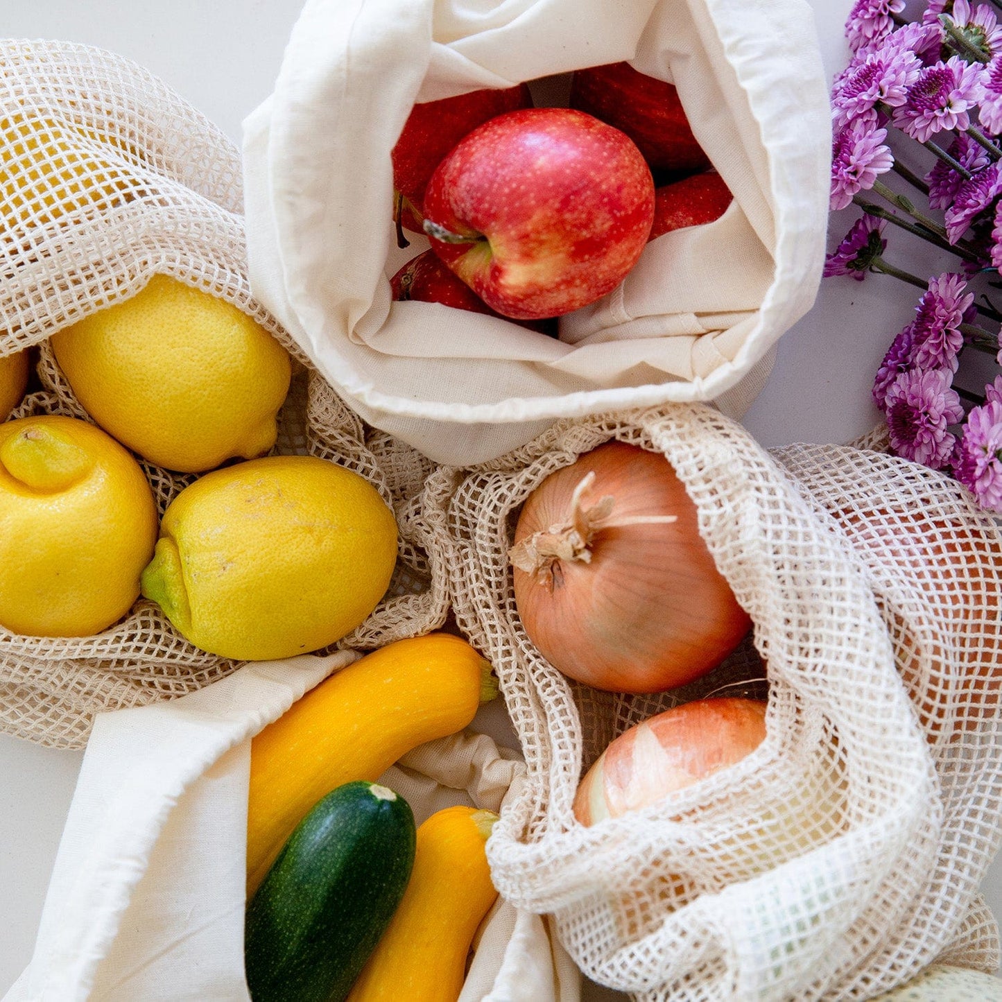 ZeroWasteStore.com's Muslin Produce Bag - Multiple Sizes holds apples, lemons, zucchini, and onions on a white surface beside purple flowers—a stylish zero waste grocery solution.
