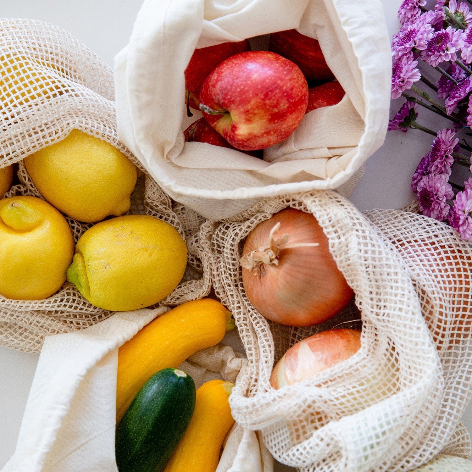 ZeroWasteStore.com's Muslin Produce Bag - Multiple Sizes holds apples, lemons, zucchini, and onions on a white surface beside purple flowers—a stylish zero waste grocery solution.