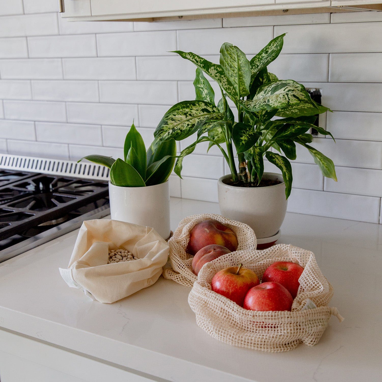 Two potted green plants, apples in a ZeroWasteStore.com Muslin Produce Bag, peaches in a Zero Waste Muslin Bag, and white beans in a cloth pouch rest on a white kitchen counter near the stove and tile backsplash.