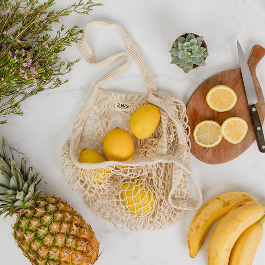 The Organic Cotton String Bag by ZeroWasteStore.com is filled with lemons and placed on a white surface, surrounded by a pineapple, bananas, fresh herbs, a succulent, and a wooden board with sliced lemons and a knife.