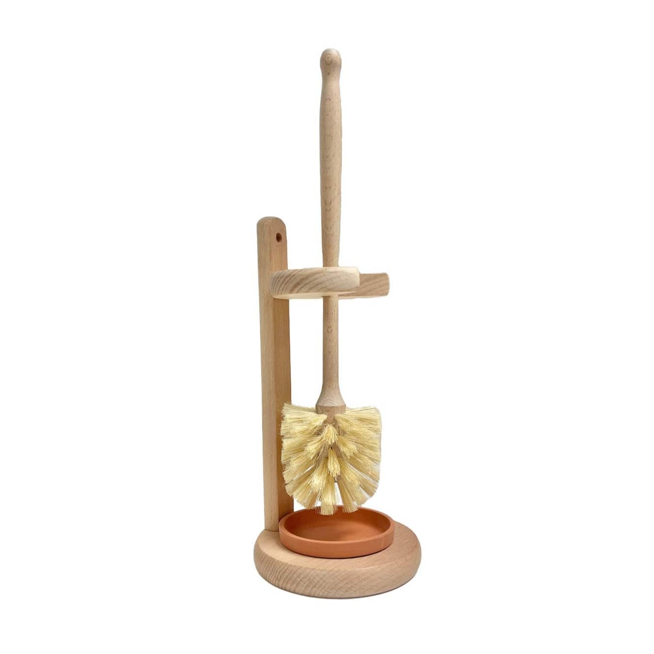 The Plastic-Free Toilet Brush from ZeroWasteStore.com features light bristles, an upright wooden handle, a matching stand, and a small round terracotta dish at the base, echoing a classic wooden toilet brush design.