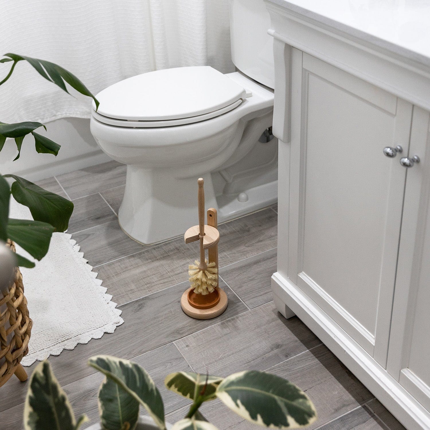 A modern bathroom with gray wood-look tile, white toilet and vanity cabinet, leafy plants, a white rug, and the Plastic-Free Toilet Brush by ZeroWasteStore.com—an eco-friendly beech wood brush—placed beside the toilet.