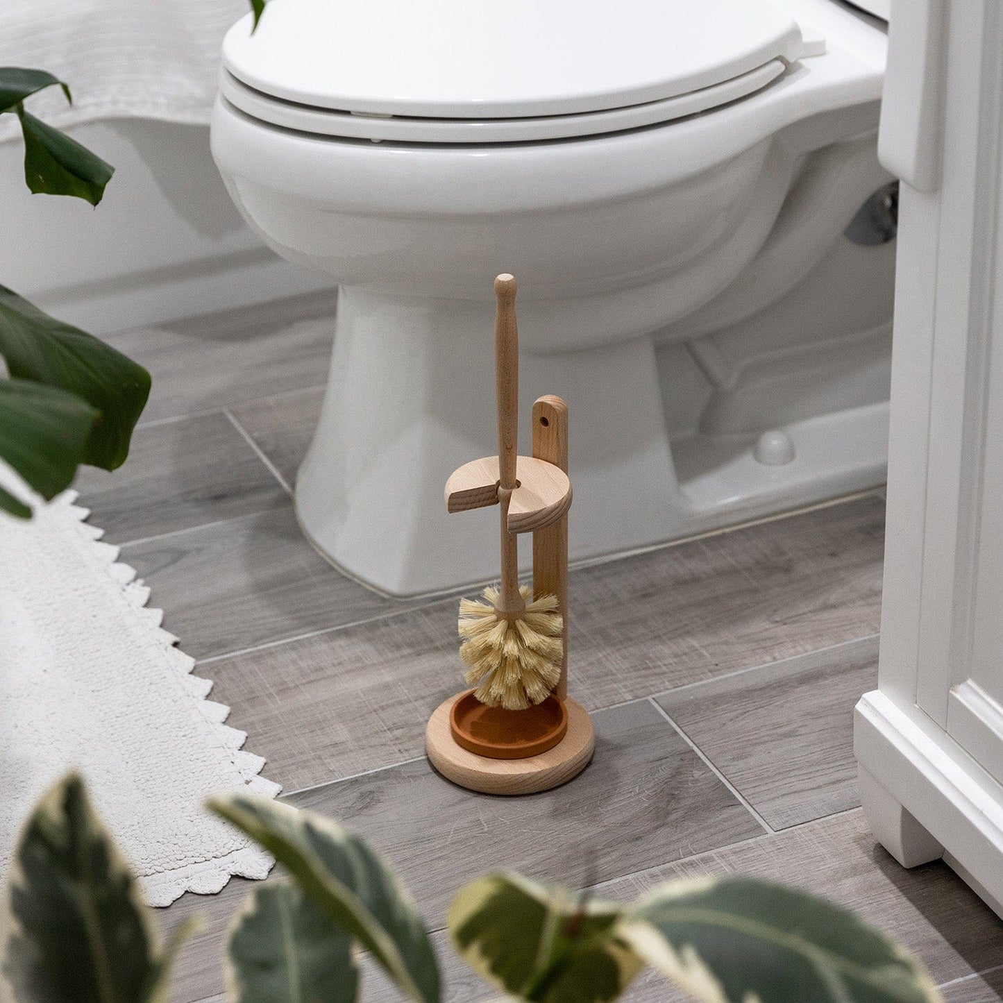 The Plastic-Free Toilet Brush by ZeroWasteStore.com sits on gray tiles near a white toilet, surrounded by green plants and a white rug, adding an eco-friendly and natural touch to your bathroom.