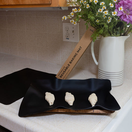 A ZeroWasteStore.com Silicone Baking Mat with three unbaked croissants rests on a tray on the kitchen counter, next to a labeled box for the mat and a pitcher of flowers.