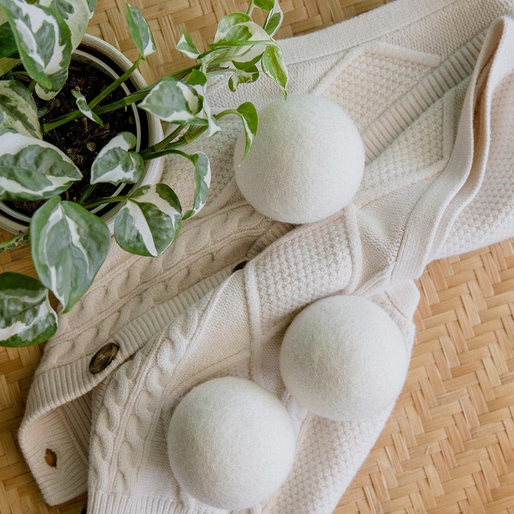 Three ZeroWasteStore.com Reusable Wool Dryer Balls are arranged on a cream knitted sweater with wooden buttons next to a green and white potted plant, all placed neatly atop a woven mat.
