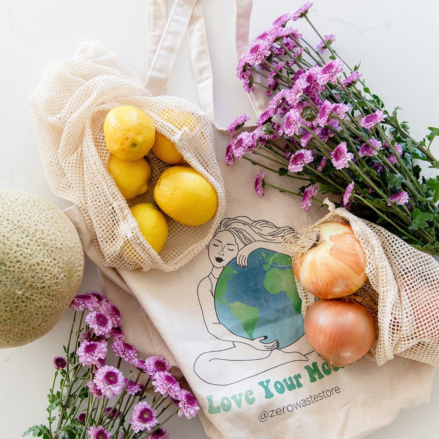 A reusable organic cotton tote bag with “Love Your Mother” art sits beside ZeroWasteStore.com Mesh Produce Bags, filled with lemons, onions, cantaloupe, and flowers—perfect for a sustainable zero waste lifestyle.