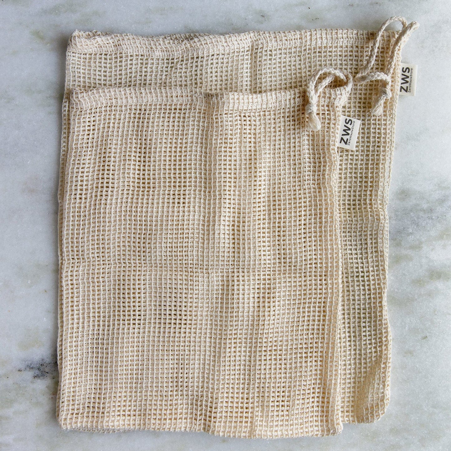 Three beige ZeroWasteStore.com Mesh Produce Bags in various sizes, made from organic cotton with drawstrings and ZWS labels, are laid flat on a light marble surface—perfect for a zero-waste lifestyle.