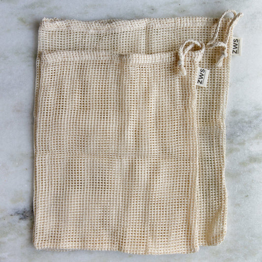 Three beige ZeroWasteStore.com Mesh Produce Bags in various sizes, made from organic cotton with drawstrings and ZWS labels, are laid flat on a light marble surface—perfect for a zero-waste lifestyle.