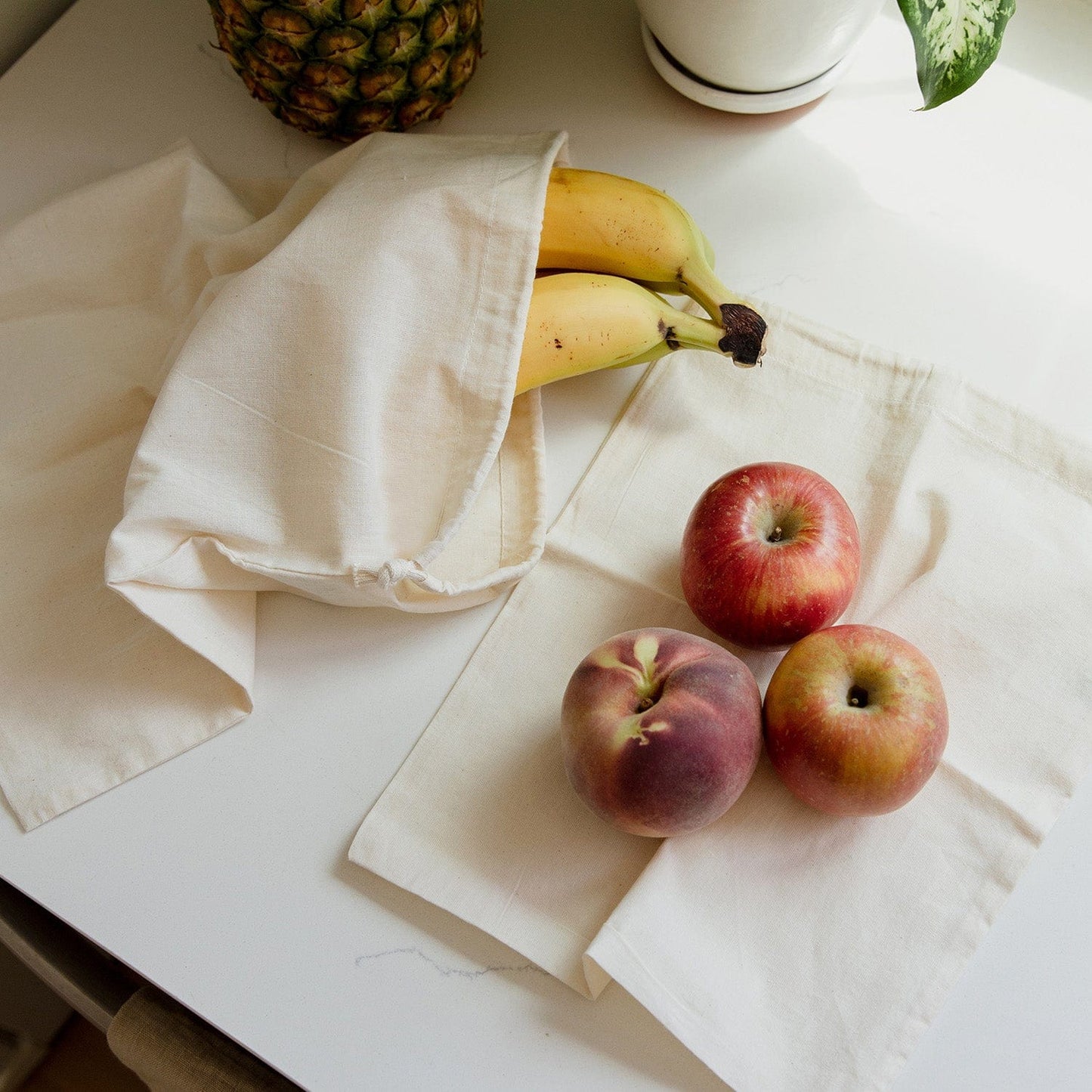 Bananas in a ZeroWasteStore.com Muslin Produce Bag, with two apples and a peach on cream cloths atop a white surface. A pineapple and potted plant are partly visible in the background.