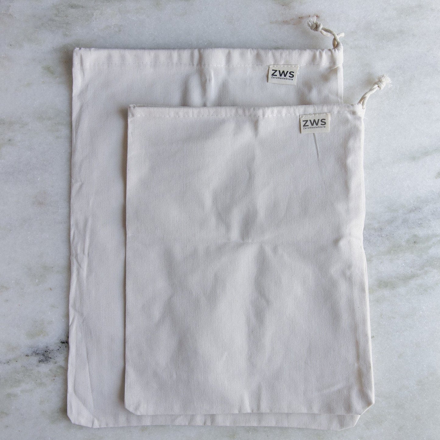 Two off-white Muslin Produce Bags from ZeroWasteStore.com, available in multiple sizes and featuring a ZWS tag, rest on a marble surface—perfect as organic, reusable bags for a zero waste grocery lifestyle.