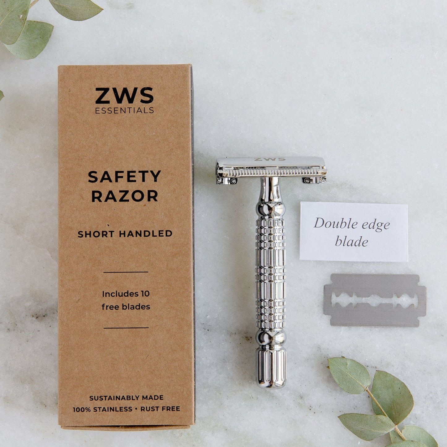 The ZeroWasteStore.com Zero Waste Safety Razor, a unisex metal single-blade razor with 10 free blades, rests on white marble beside its branded box, a double-edge blade, and a note—all arranged among fresh green leaves.