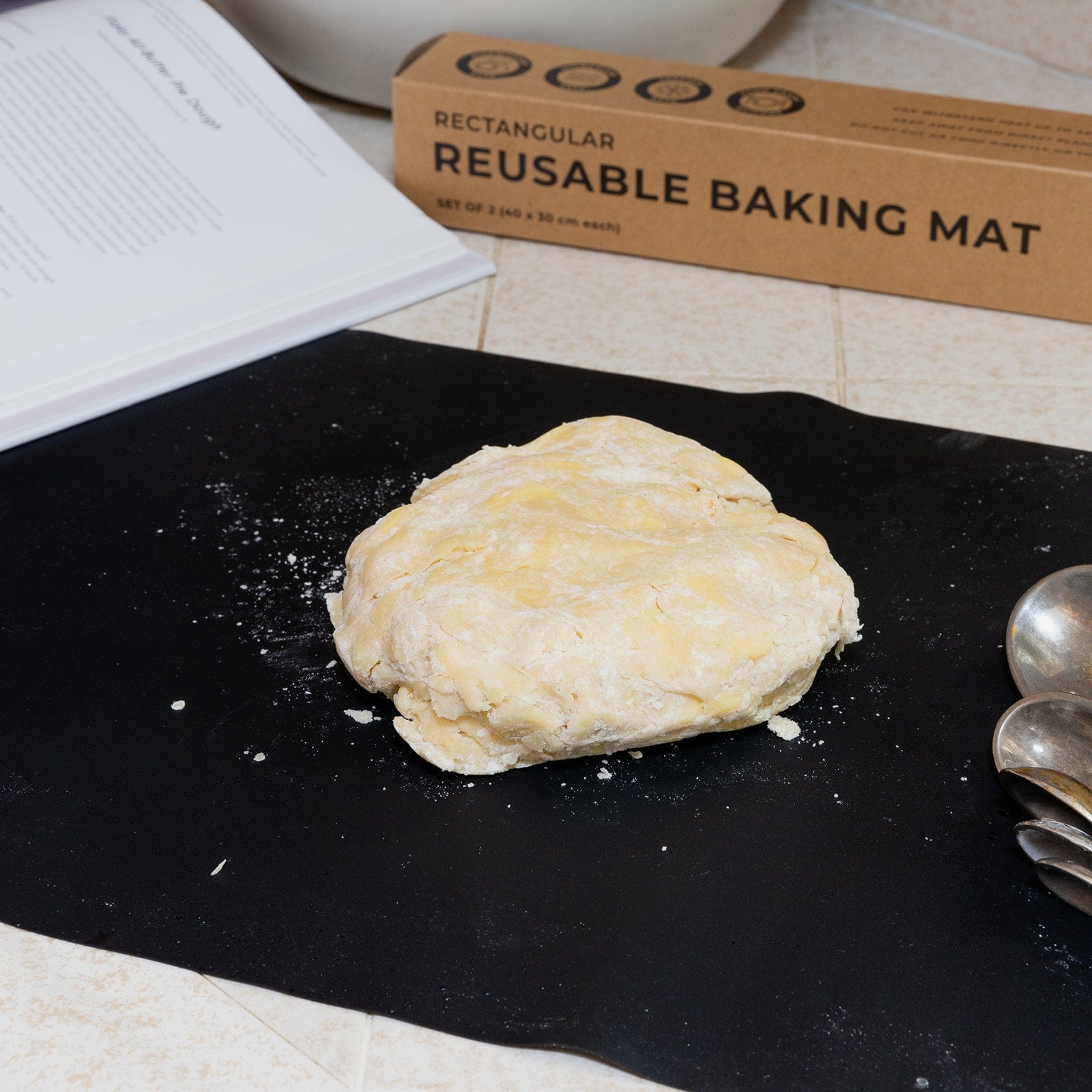 A ball of dough sits on a black ZeroWasteStore.com Silicone Baking Mat on the kitchen counter, surrounded by metal measuring spoons, a recipe book, and the product’s box labeled “Rectangular Reusable Baking Mat.”.