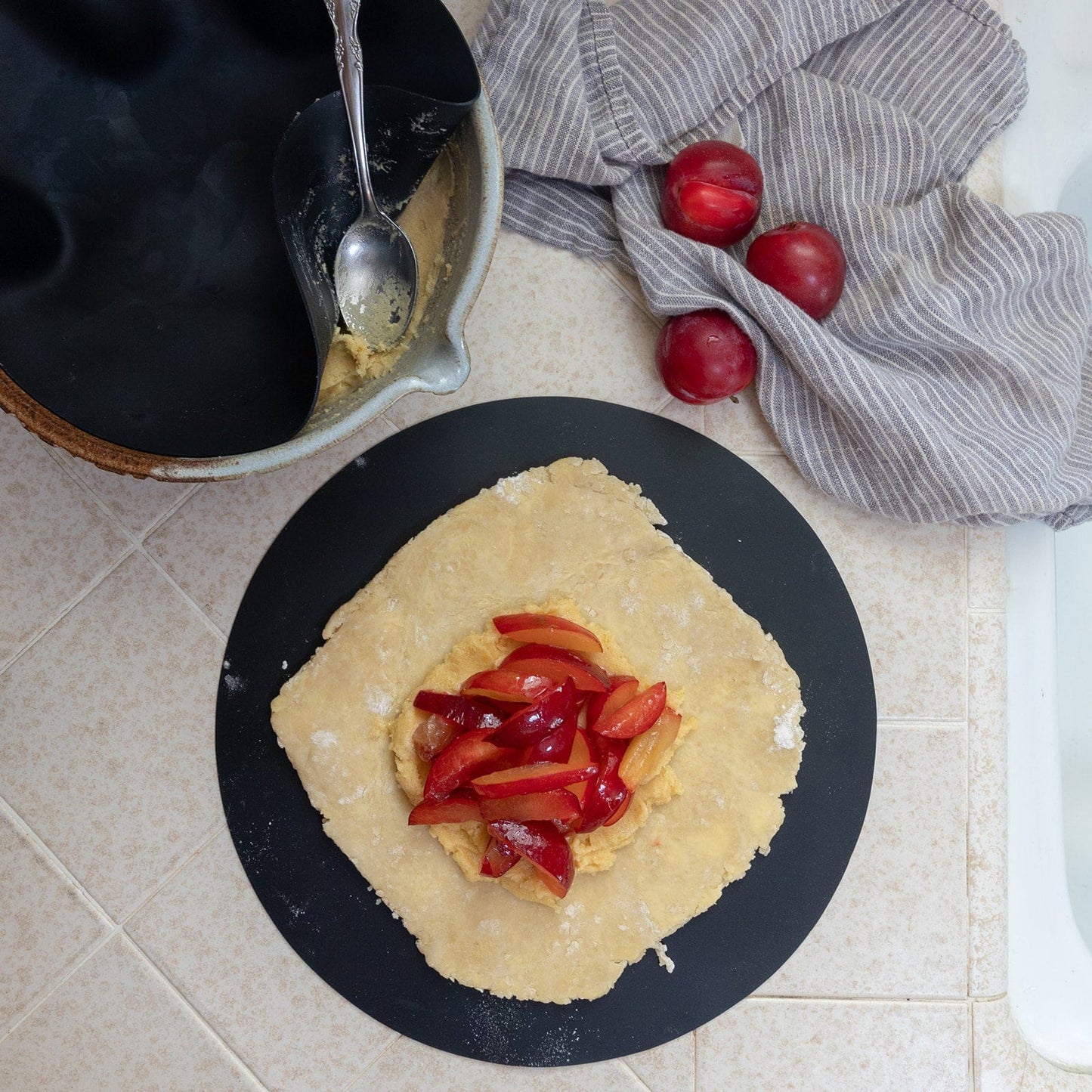 A square piece of dough on the ZeroWasteStore.com Silicone Baking Mat holds sliced red plums at the center. Nearby are whole plums, a gray striped towel, a mixing bowl with dough remnants, and a spoon on the tiled counter.