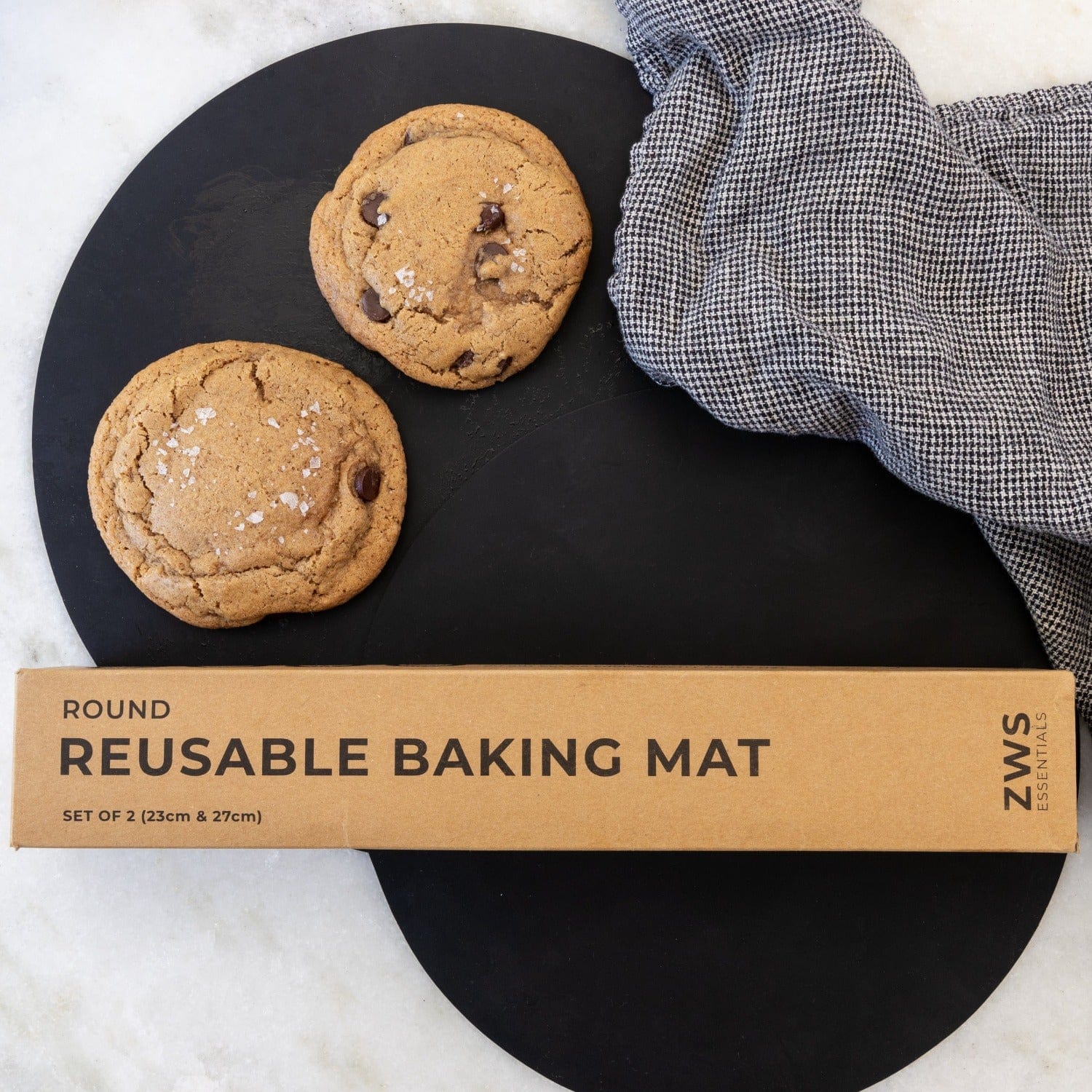 Two chocolate chip cookies sit on a black surface beside a textured gray cloth. In front, a brown box labeled Silicone Baking Mat from ZeroWasteStore.com promotes their durable, non-stick silicone mat for easy baking.