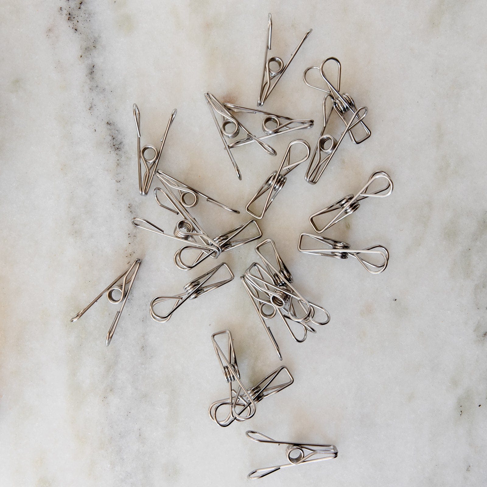 ZeroWasteStore.com Stainless Steel Clips—Laundry Pegs, Clothespins, Multipurpose Clips (20 Pack)—are scattered on a marble surface, showcasing their simple, eco-friendly design as a stylish plastic-free alternative for everyday use.