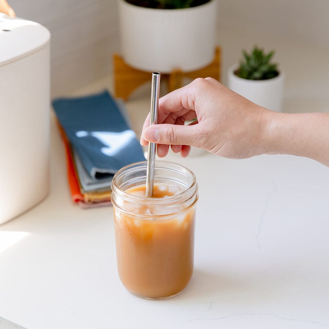 A hand stirs iced coffee in a mason jar using a ZeroWasteStore.com Stainless Steel Straw (Straight, Bent, or Boba Style) on a white countertop. Folded cloth napkins and potted plants add to the eco-friendly, plastic-free scene.