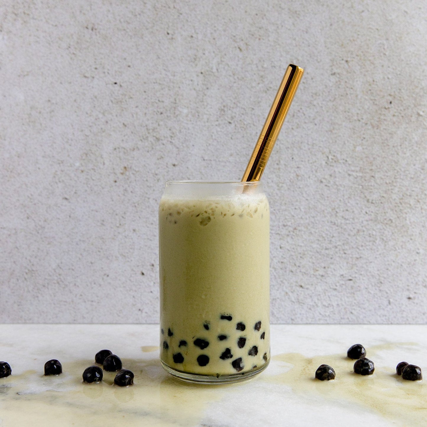 A glass of iced bubble tea with black tapioca pearls, foam on top, and a ZeroWasteStore.com Stainless Steel Straw - Boba Style. Extra tapioca pearls rest on the white marble surface.