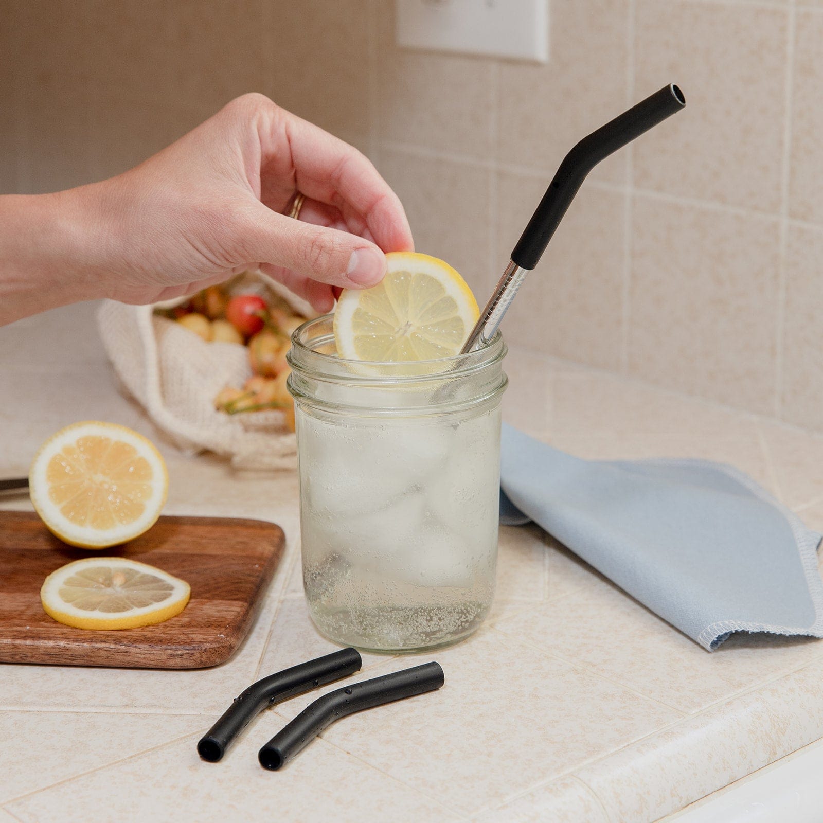 A hand adds a lemon slice to iced water in a mason jar with a stainless steel straw fitted with a ZeroWasteStore.com Silicone Straw Tip. On the counter, two extra 100% silicone tips from the Silicone Straw Tip Pack and half a lemon sit on a wooden board.
