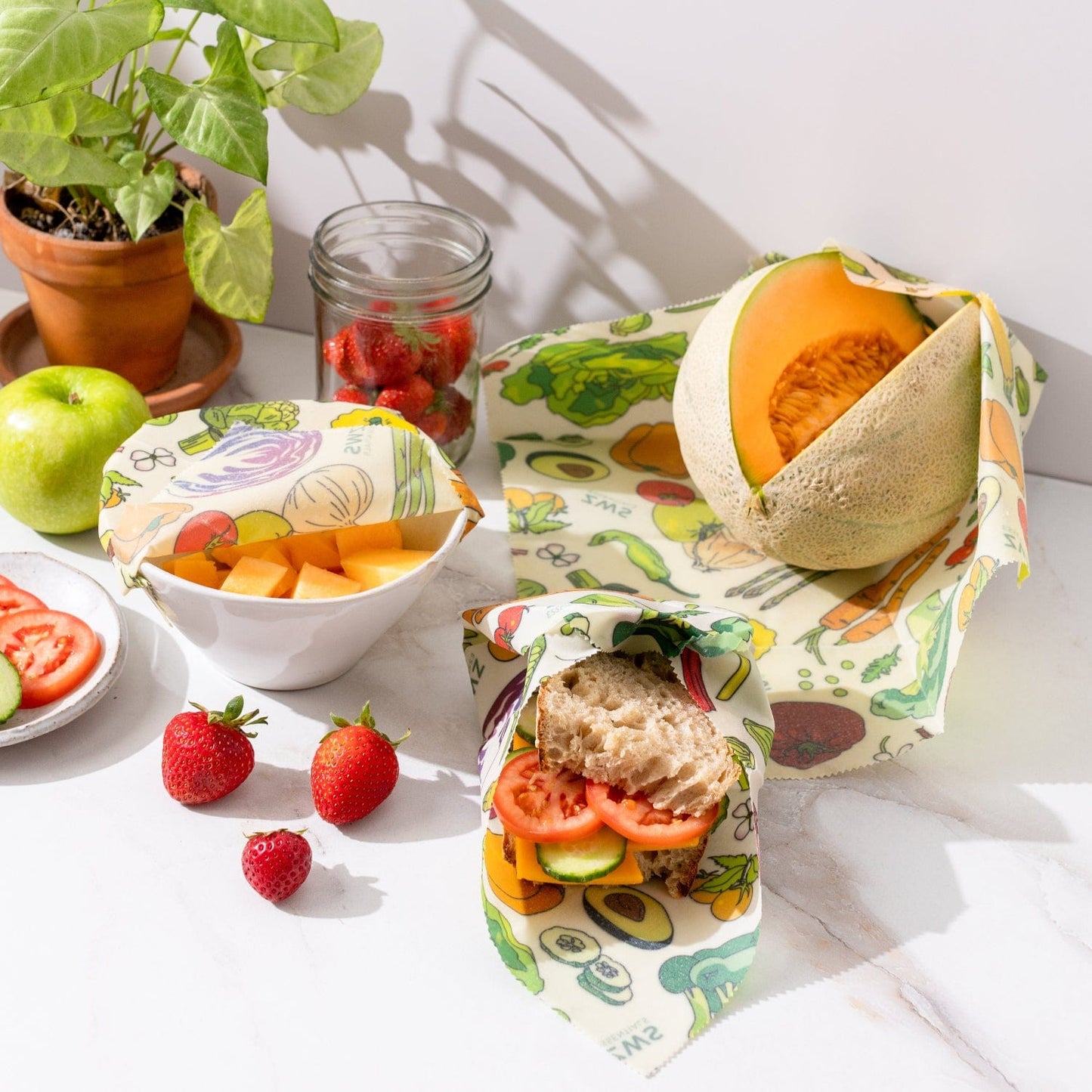 ZeroWasteStore.com’s Beeswax Wraps – Plastic-Free Food Storage cover a sandwich, bowl, and cantaloupe on a white surface with strawberries, apple, tomato slices, a plant, and a jar of strawberries nearby.
