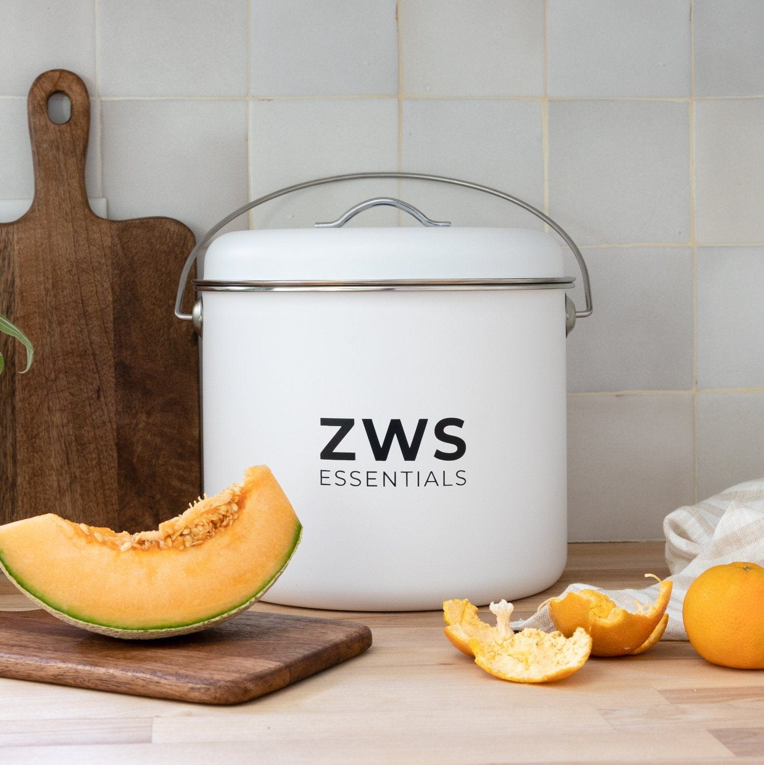 A ZeroWasteStore.com Countertop Compost Bin sits on a kitchen counter with a cantaloupe slice, orange peels, a mandarin orange, and a wooden cutting board nearby. Ideal for convenient composting. Tiled wall in the background.