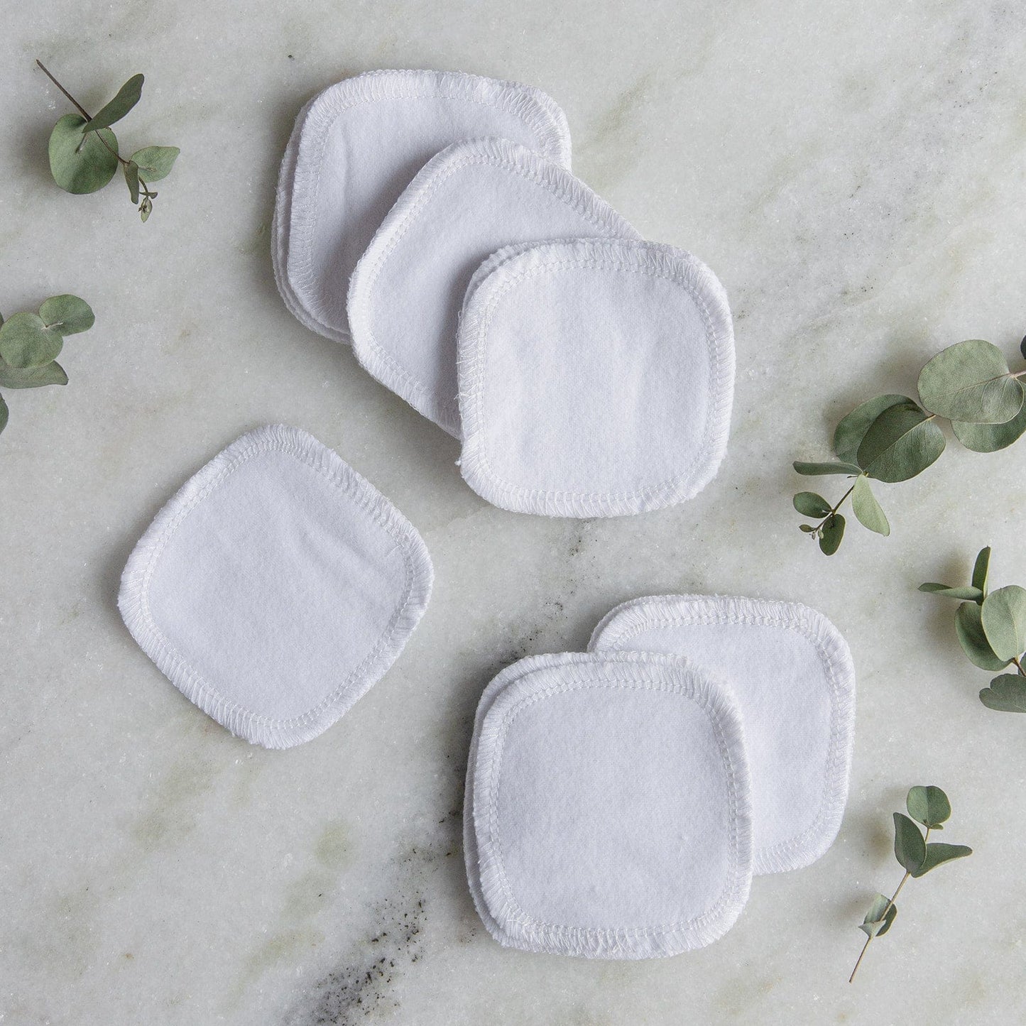 Seven Reusable Cotton Rounds from ZeroWasteStore.com, crafted from organic cotton, are displayed on a light marble surface alongside small eucalyptus sprigs.