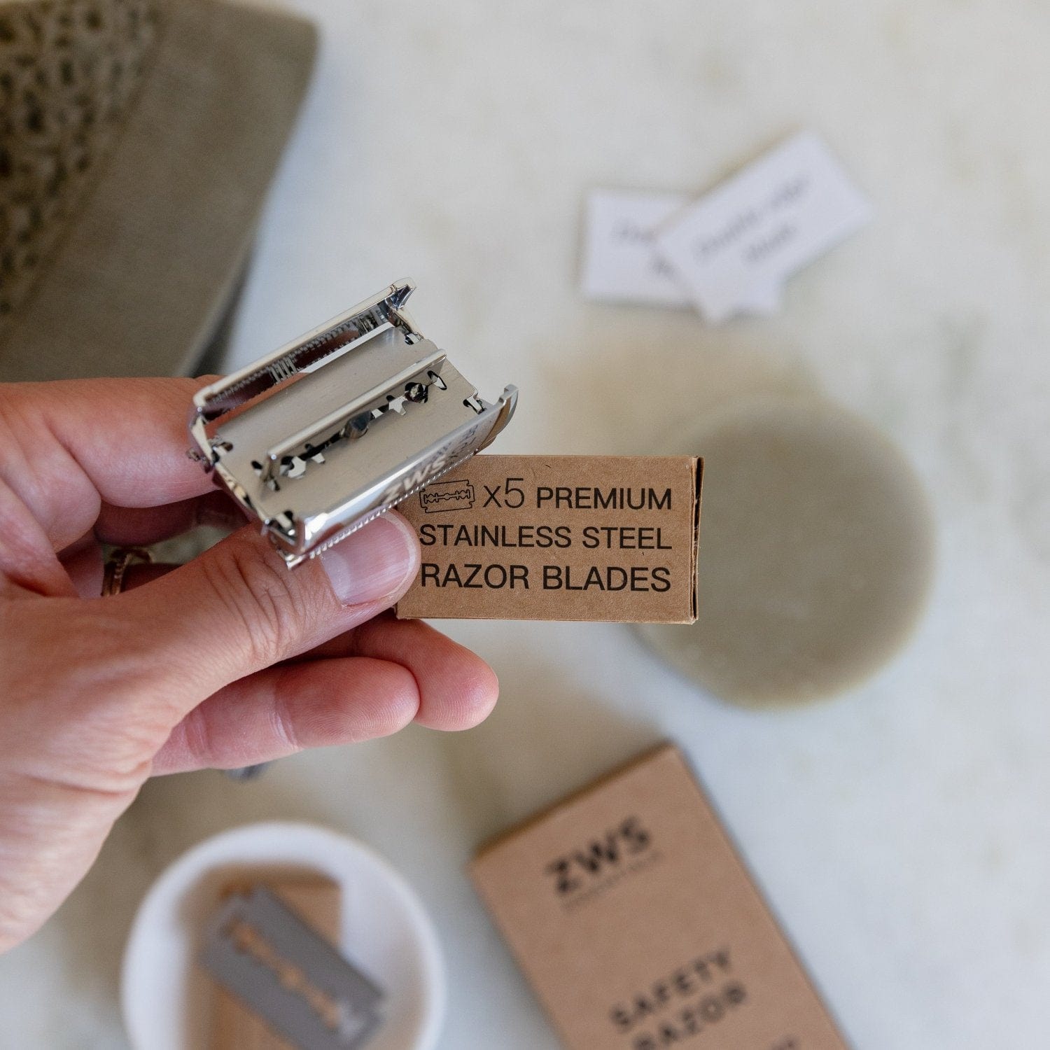 A hand holds an open Zero Waste Razor and a ZeroWasteStore.com pack labeled “Zero Waste Razor Replacement Blades.” In the background are a soap bar, box, and paper items on a light surface.