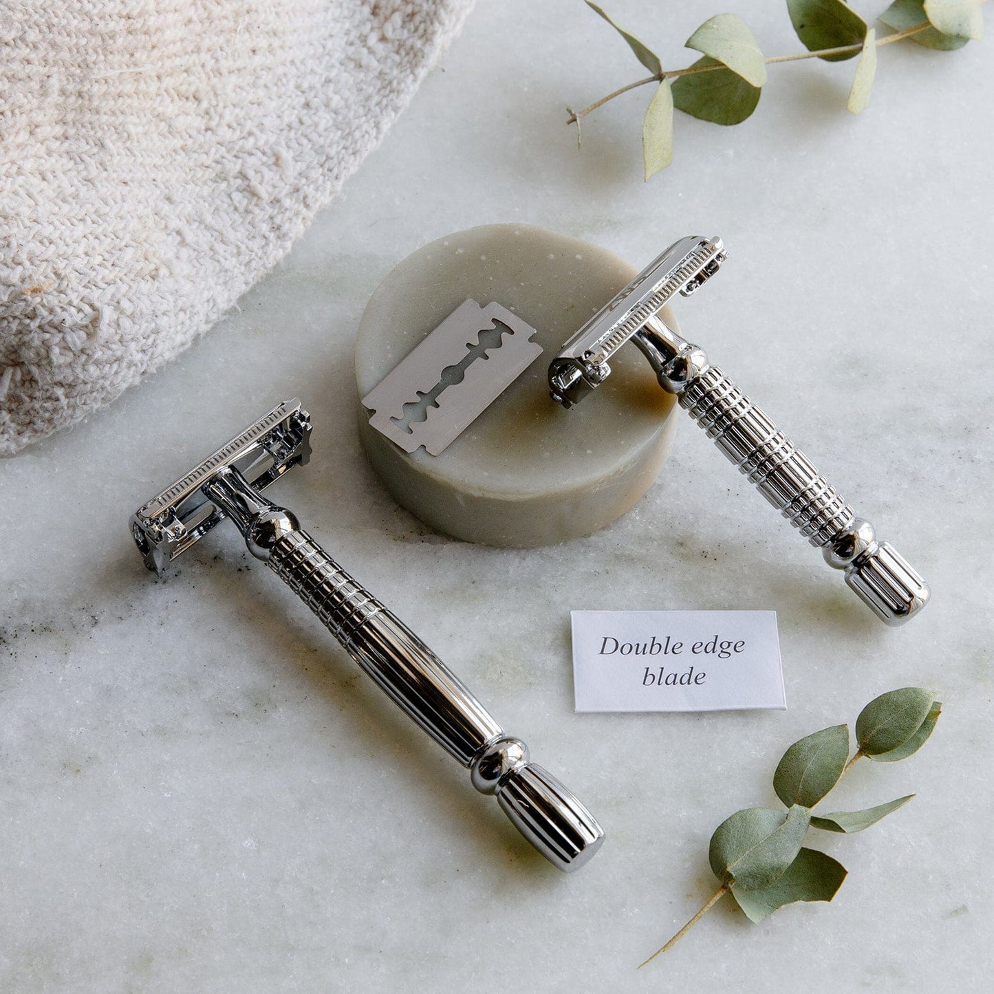 Two Zero Waste Safety Razors by ZeroWasteStore.com with 10 free blades, a round soap bar, towel, and green leaves rest on marble. A label highlights: “Double edge blade—ideal for zero plastic waste grooming.”.