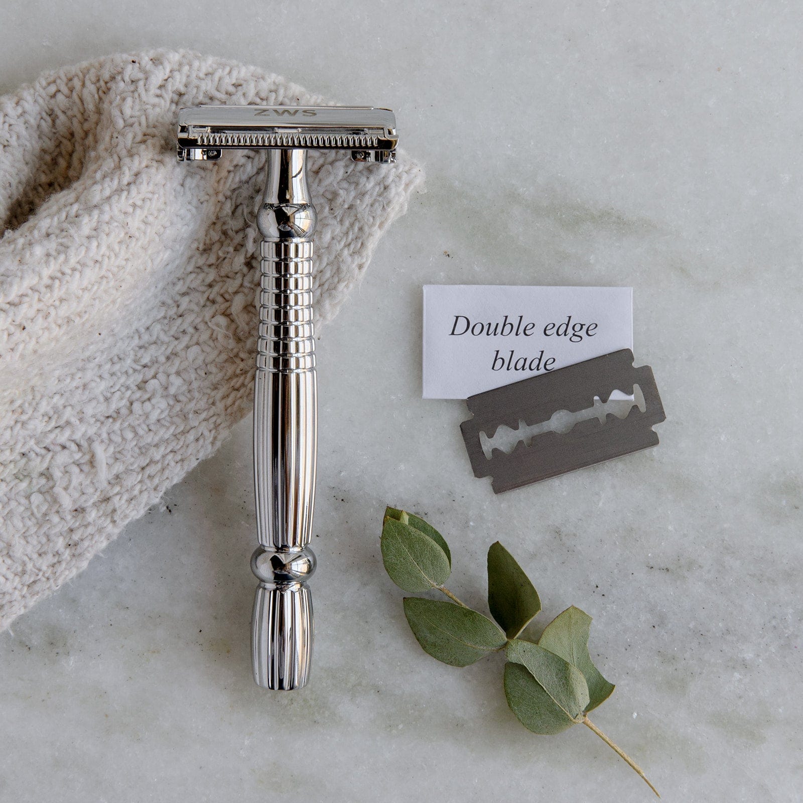 A ZeroWasteStore.com Zero Waste Safety Razor with a single blade and 10 free blades rests on textured cloth by a Double edge blade pack and green leaves, all displayed on marble for sustainable, plastic-free grooming.