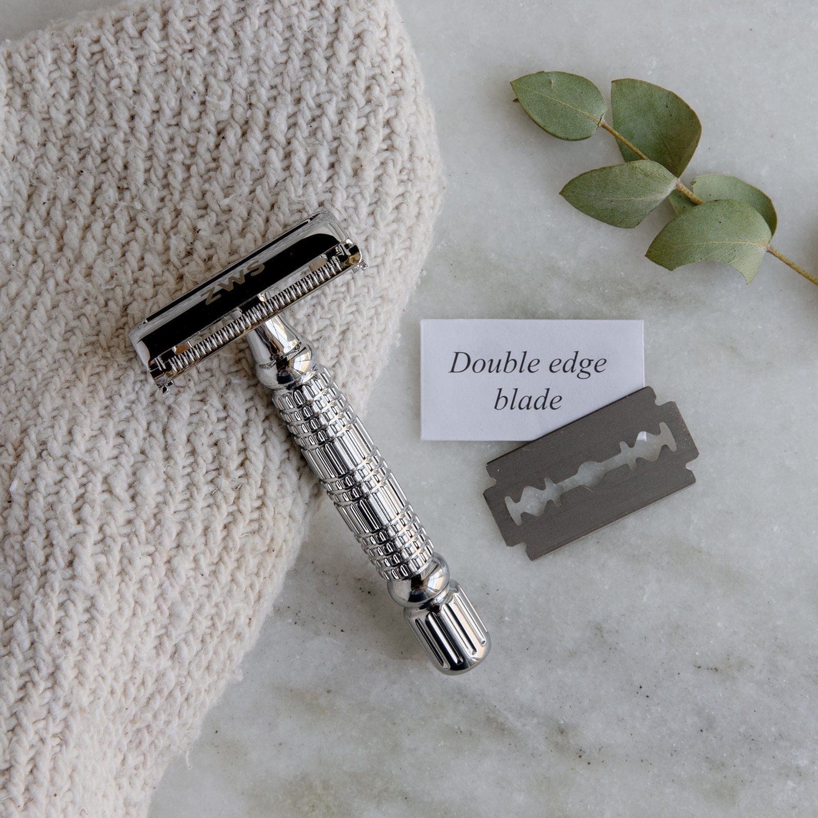 The ZeroWasteStore.com Zero Waste Safety Razor—unisex, single blade, with 10 free blades—rests on a white cloth with a razor blade, note saying Double edge blade, and eucalyptus sprig atop marble for plastic-free grooming.