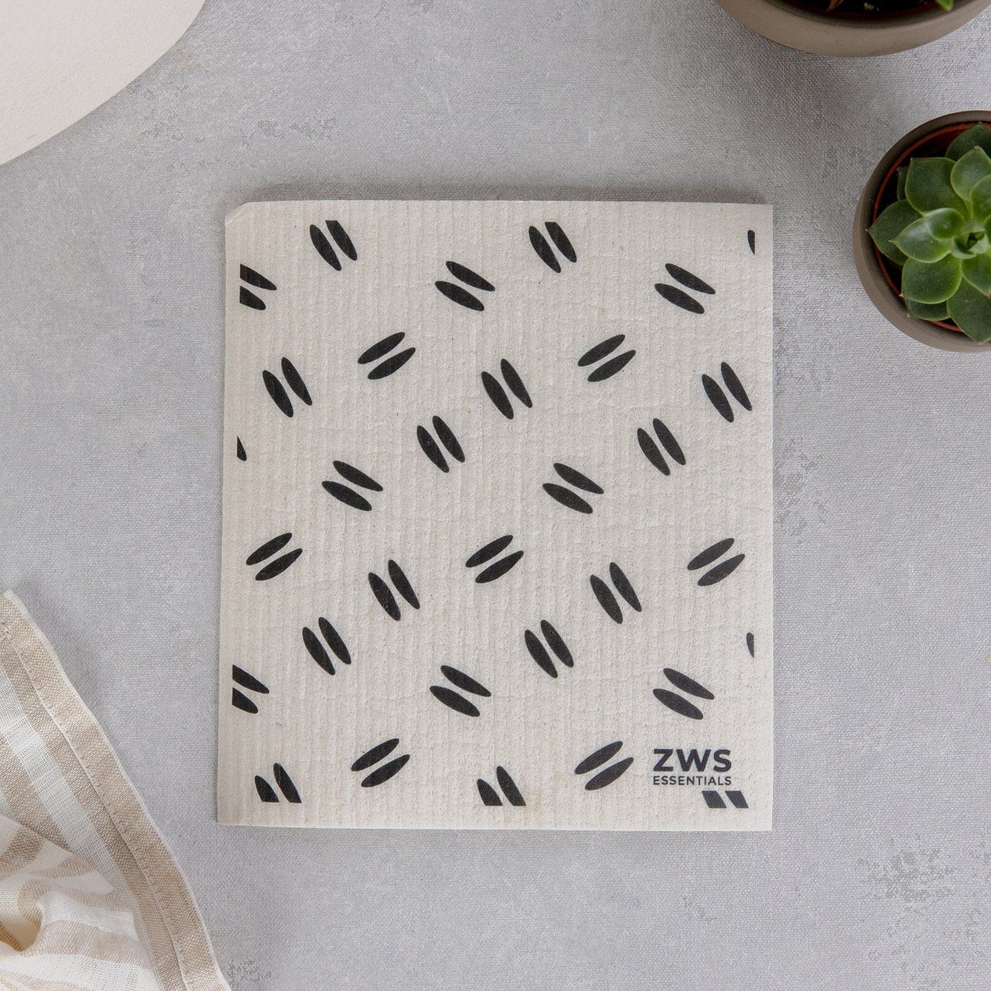 A beige Swedish Dish Cloth from ZeroWasteStore.com with black ovals and "ZWS ESSENTIALS" text sits on a light gray surface, beside a succulent, striped cloth, and round object—a perfect eco-friendly paper towel alternative.