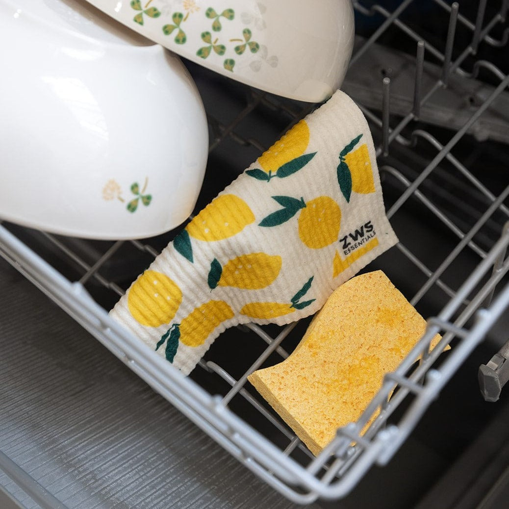 A dishwasher rack holds clean dishes, a yellow sponge, and a Swedish Dish Cloth from ZeroWasteStore.com with a lemon pattern—an eco-friendly paper towel replacement.