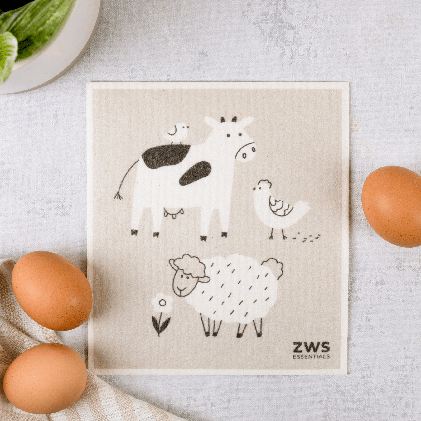 A beige Swedish Dish Cloth from ZeroWasteStore.com with black and white animal illustrations rests on a light surface near three brown eggs, a striped towel, and a potted plant.