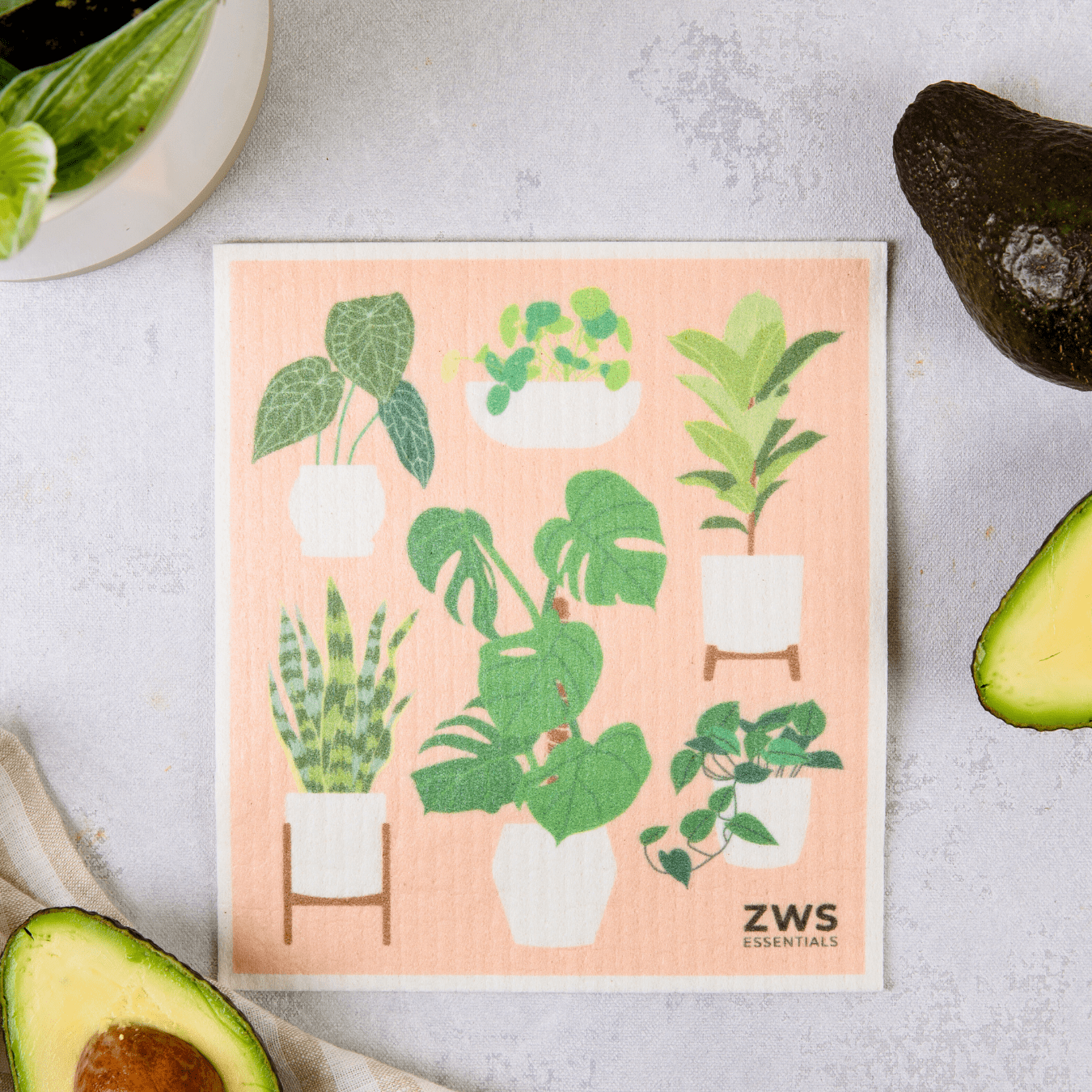 A square Swedish Dish Cloth from ZeroWasteStore.com, featuring six potted green houseplants, rests on a light surface among avocados, leafy greens, and part of a striped towel—an eco-friendly paper towel replacement.