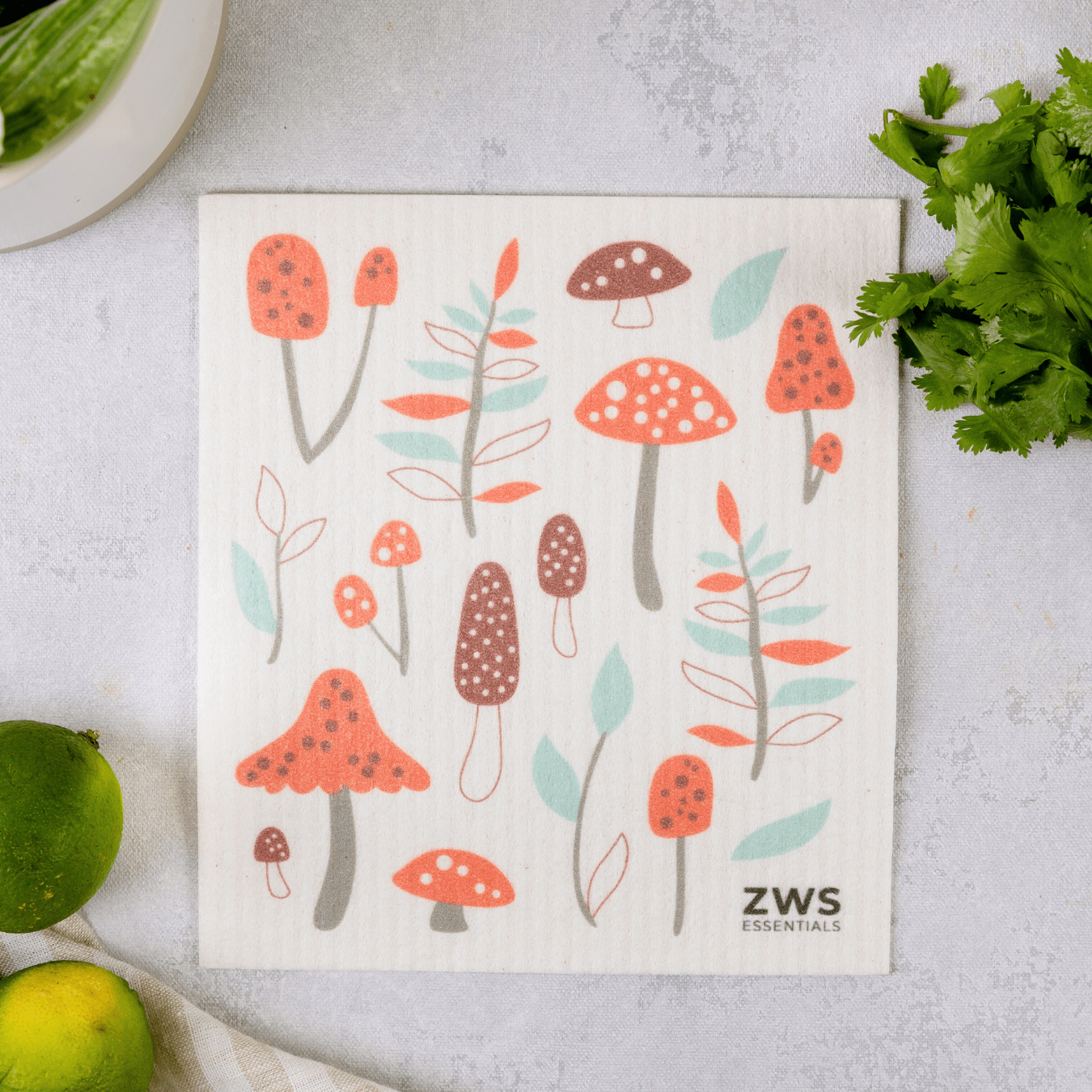 A white Swedish Dish Cloth from ZeroWasteStore.com, featuring red mushrooms and green leaves, rests on a light counter with cilantro, limes, and a bowl—a charming paper towel alternative for your sustainable kitchen.