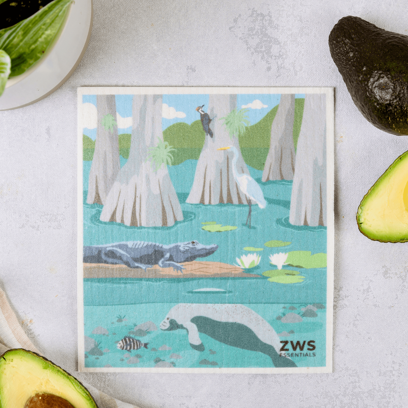 A rectangular Swedish Dish Cloth by ZeroWasteStore.com, illustrated with a wetland scene of an alligator, heron, fish, and manatee among cypress trees and lily pads, serves as a charming paper towel replacement near avocado halves and a potted plant.